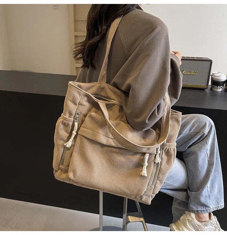 A person is holding a beige corduroy shoulder bag with a zipper on a dark surface.