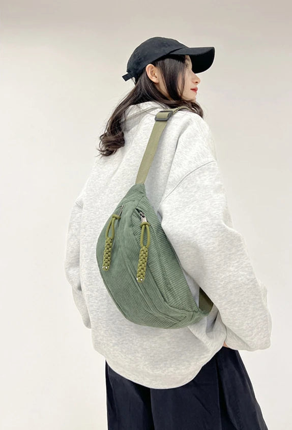 A woman wearing a light gray hoodie and black hat carrying an olive green corduroy waist pack.