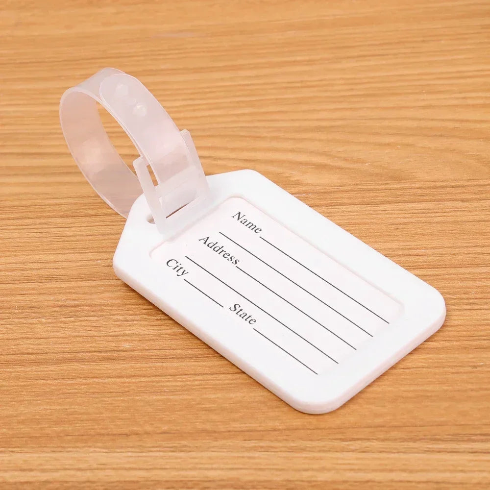 A white plastic luggage tag with a clear strap and lines for writing name, address, and state, resting on a wooden surface.