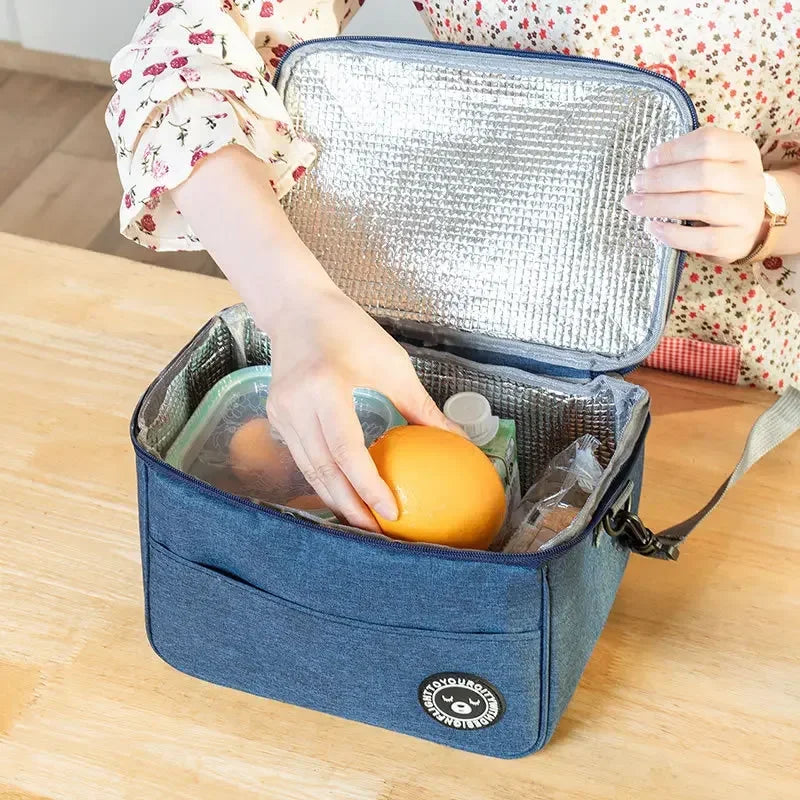A hand is opening a blue insulated portable lunch bag with a silver foil lid, revealing food inside.