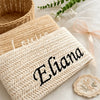 A beige handwoven straw makeup bag with black lettering rests on a white surface next to a woven basket.