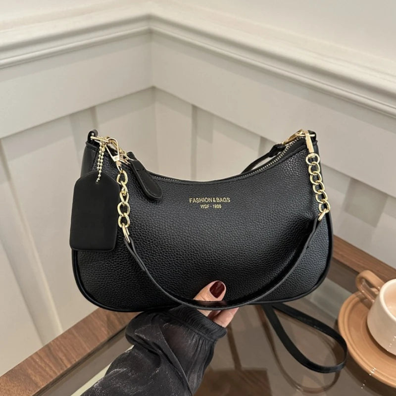 A black leather shoulder bag with a gold chain detail is held by a hand against a light background.