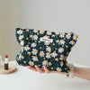 A hand holds a floral canvas makeup bag featuring white and yellow daisies on a black background.