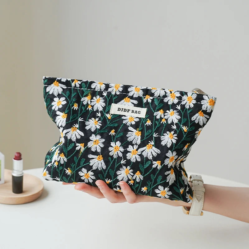 A hand holds a floral canvas makeup bag featuring white and yellow daisies on a black background.