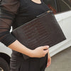 A woman holds a dark brown alligator print clutch bag against a white car.