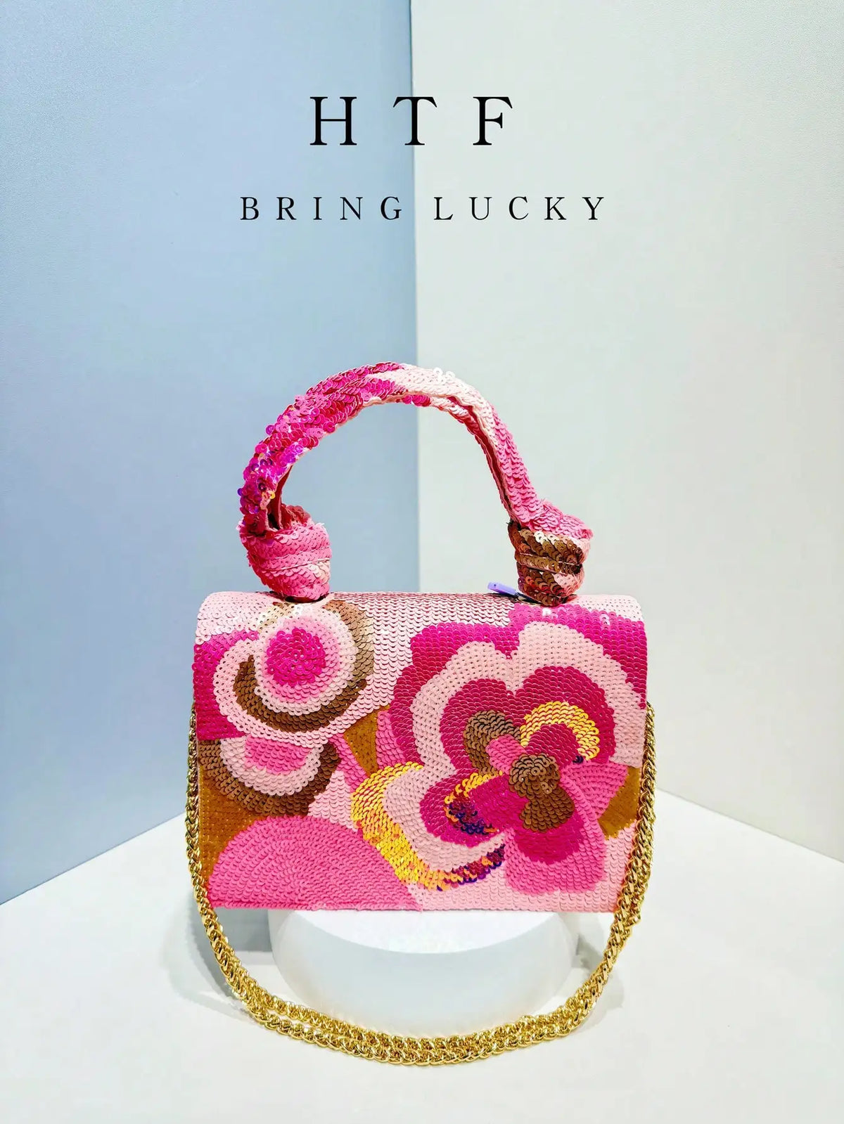 A pink floral clutch bag with gold sequins and embroidery is displayed against a light background.