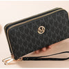 A black patterned wallet with gold zipper details and a black strap is shown being held by a hand.