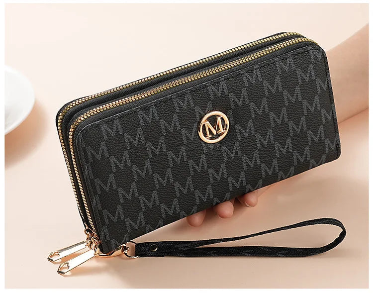 A black patterned wallet with gold zipper details and a black strap is shown being held by a hand.