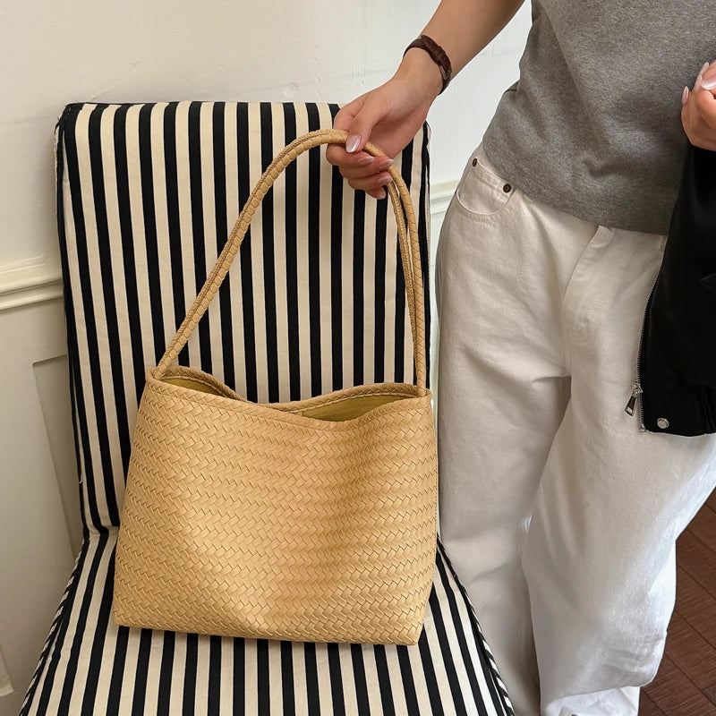 A person holds a light tan woven bucket shoulder bag with a rope handle against a black and white striped background.