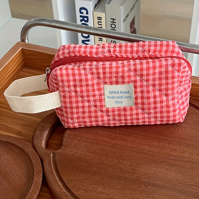 A red and white plaid cosmetic storage bag with a light beige strap resting on a wooden tray.