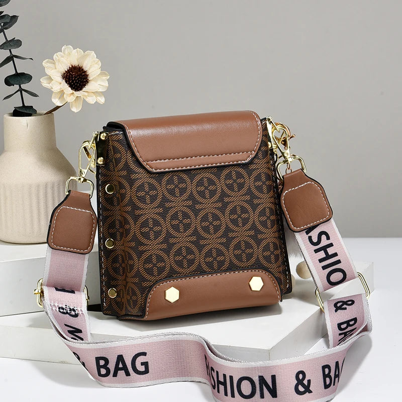 Sac bandoulière en similicuir marron avec quincaillerie dorée et sangle rose posé sur une surface blanche.