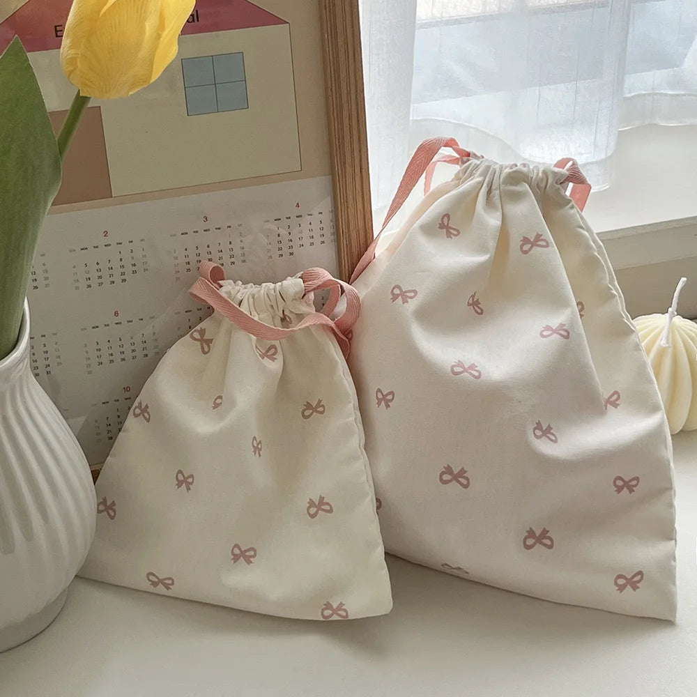 Two light pink canvas makeup bags tied with pink ribbon resting on a white surface next to a yellow flower.