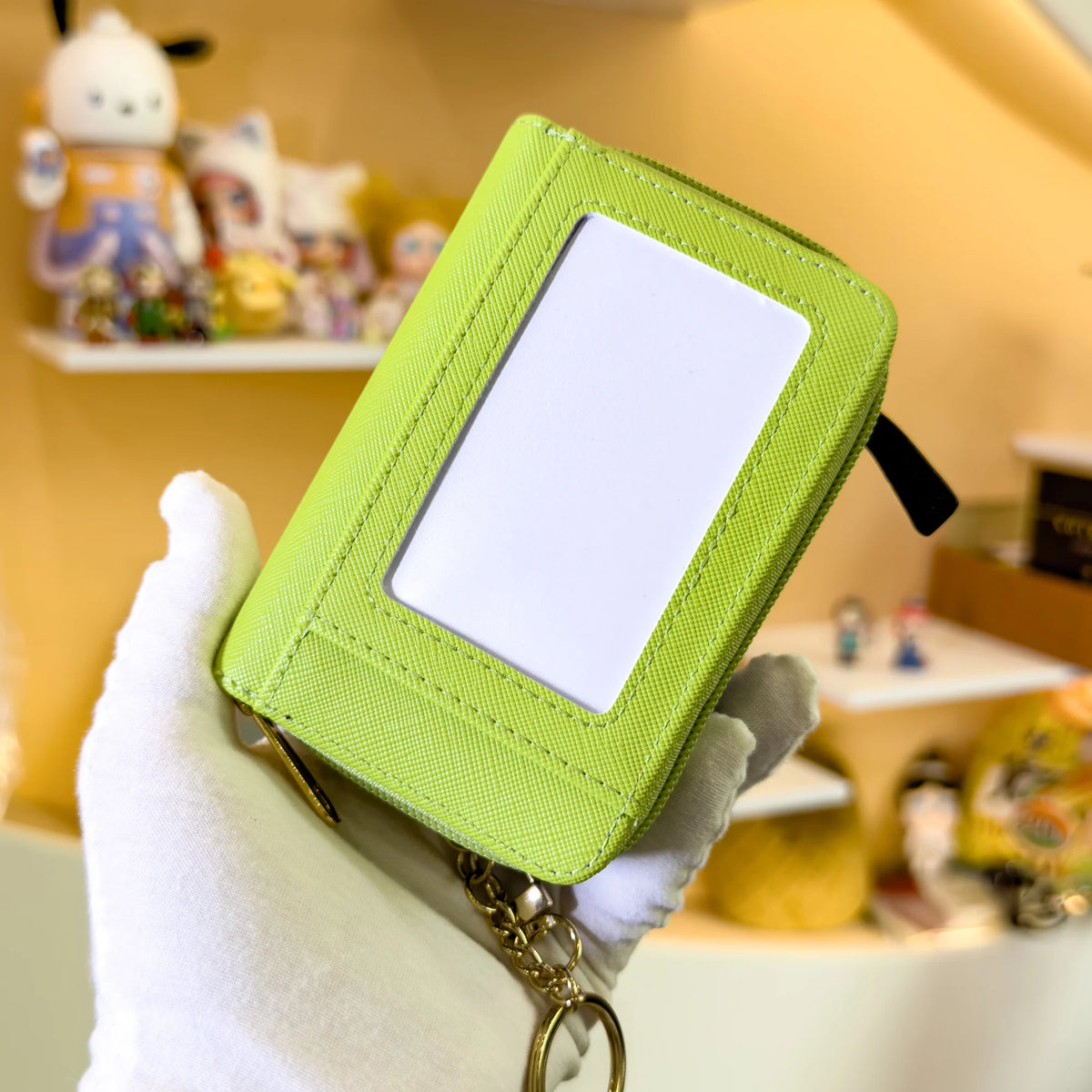 A hand holds a lime green coin purse wallet with a clear window, set against a background of small figurines.