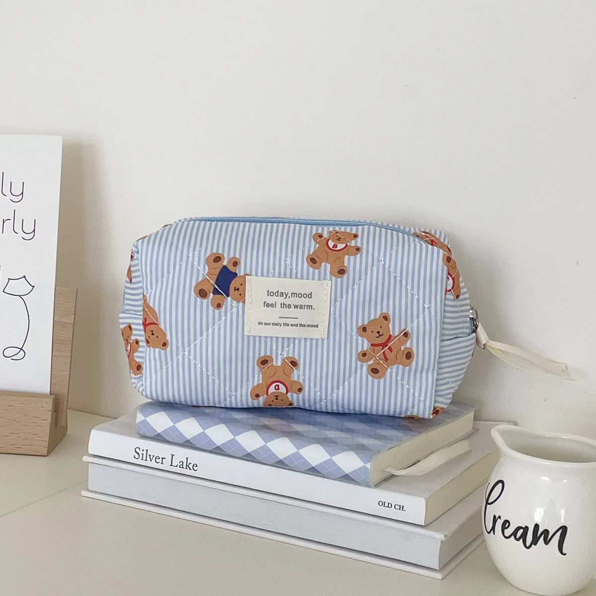 A light blue and white striped cosmetic makeup bag featuring bear designs, resting on a stack of books.
