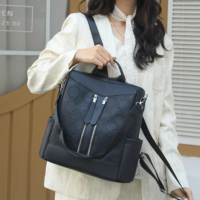 A woman is holding a black leather anti-theft backpack in front of a light beige jacket.