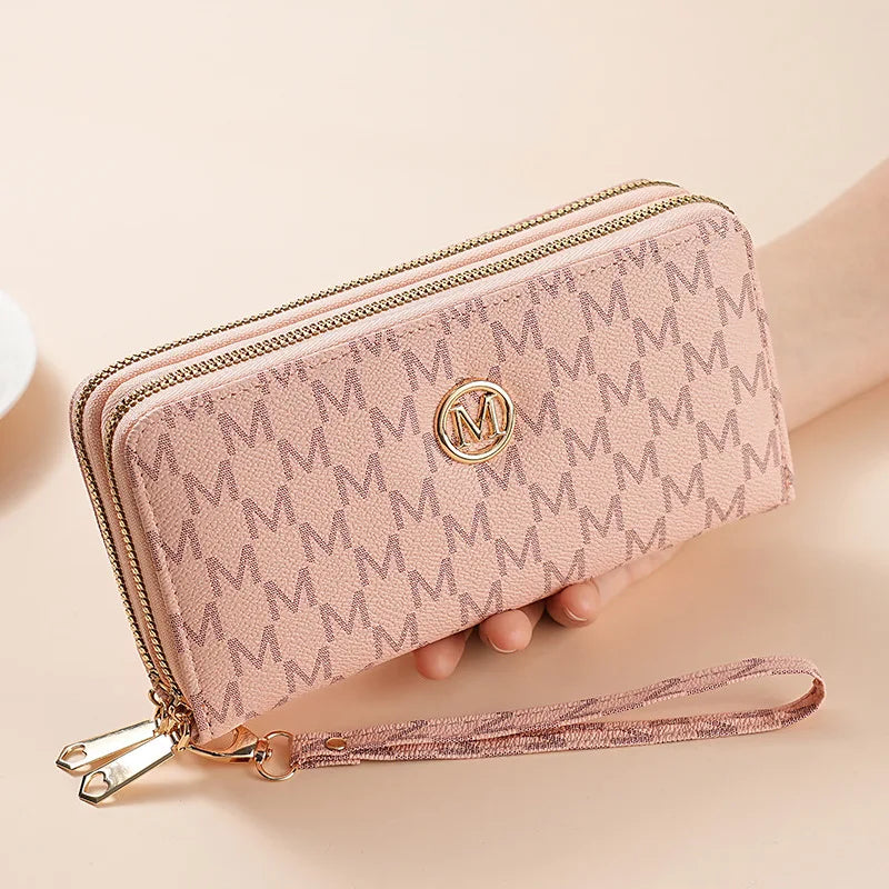 A light pink double zipper wallet with a monogram pattern and a leather strap is shown on a hand.