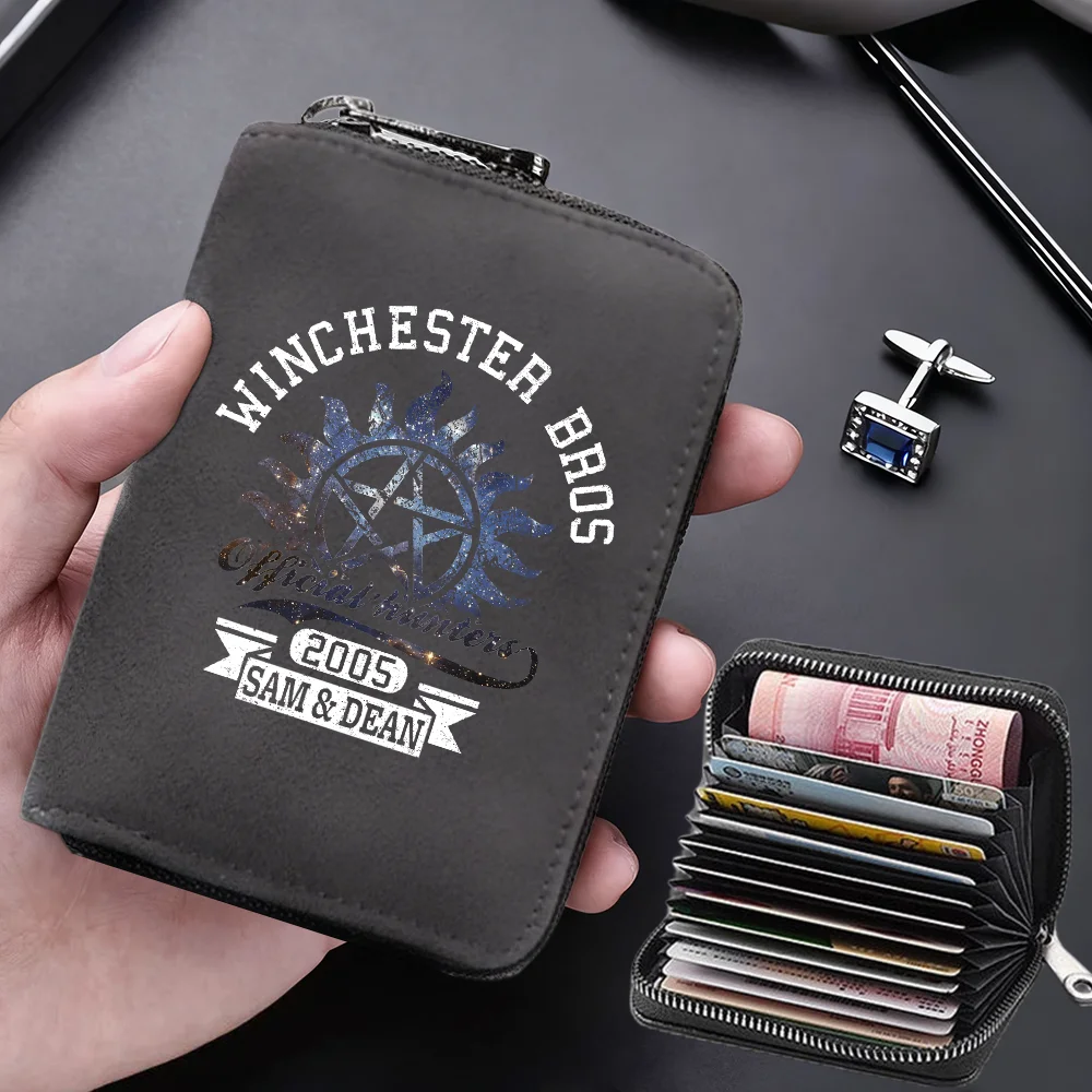 A dark gray leather wallet with a silver clasp and an engraved design is held in a hand next to a black card holder.