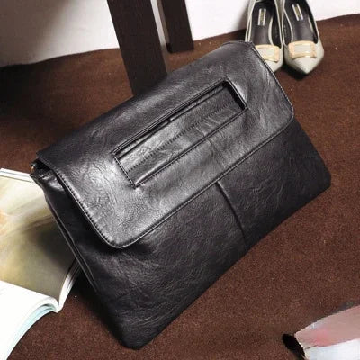 A dark gray leather crossbody clutch bag resting on a brown surface next to a pair of shoes and a book.