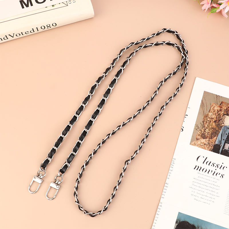A close-up of a black leather chain strap for an adjustable crossbody purse, laid diagonally on a light pink background.