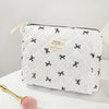A white quilted makeup bag with black floral embroidery rests on a white surface next to a gold makeup brush.