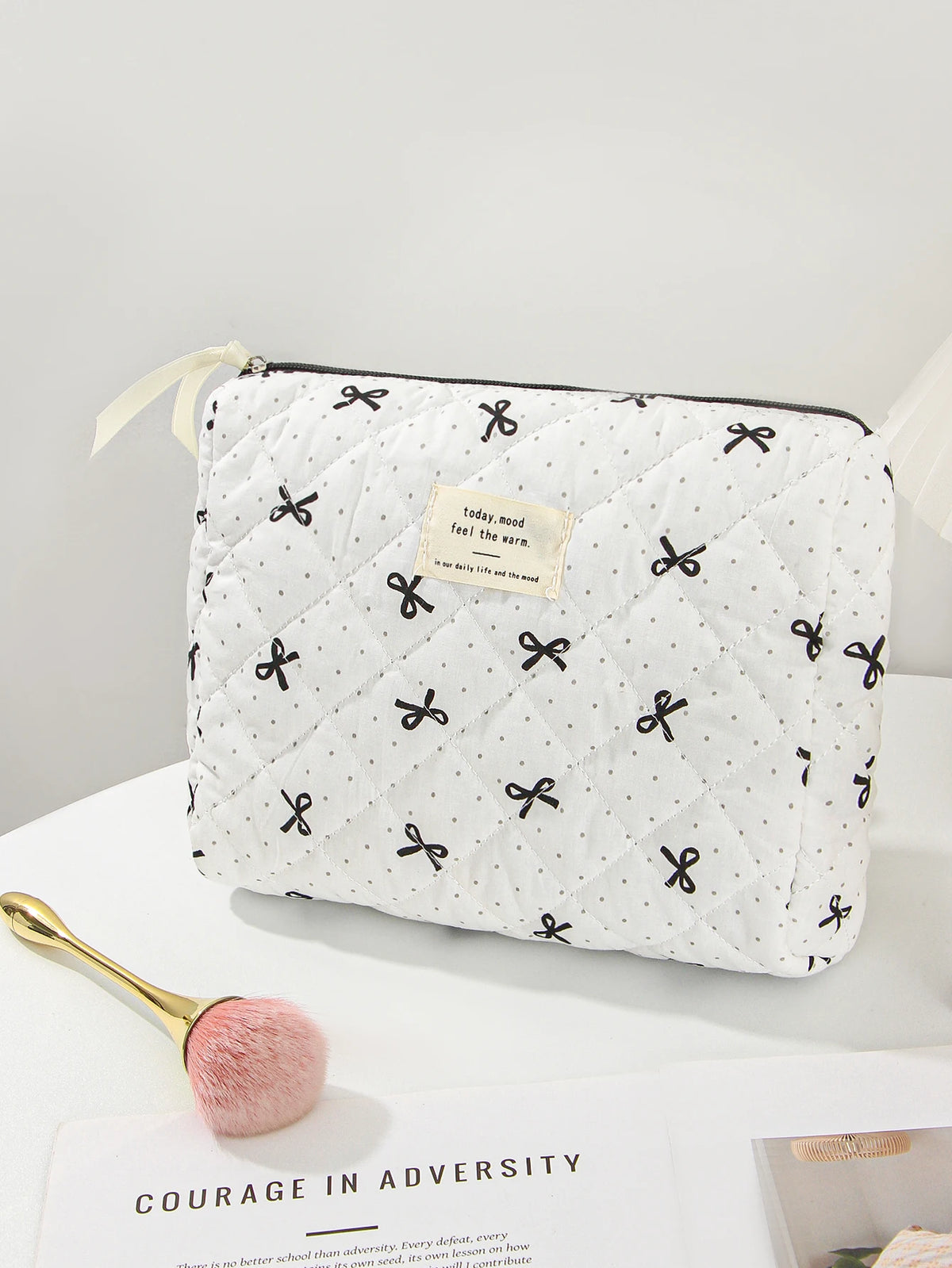 A white quilted makeup bag with black floral embroidery rests on a white surface next to a gold makeup brush.
