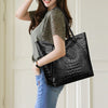 A woman holds a black crocodile print tote bag and wears a green top and blue jeans.