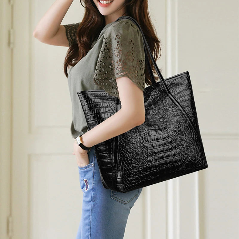 A woman holds a black crocodile print tote bag and wears a green top and blue jeans.