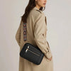 A woman wearing a beige trench coat holds a black retro square crossbody bag with an adjustable strap.