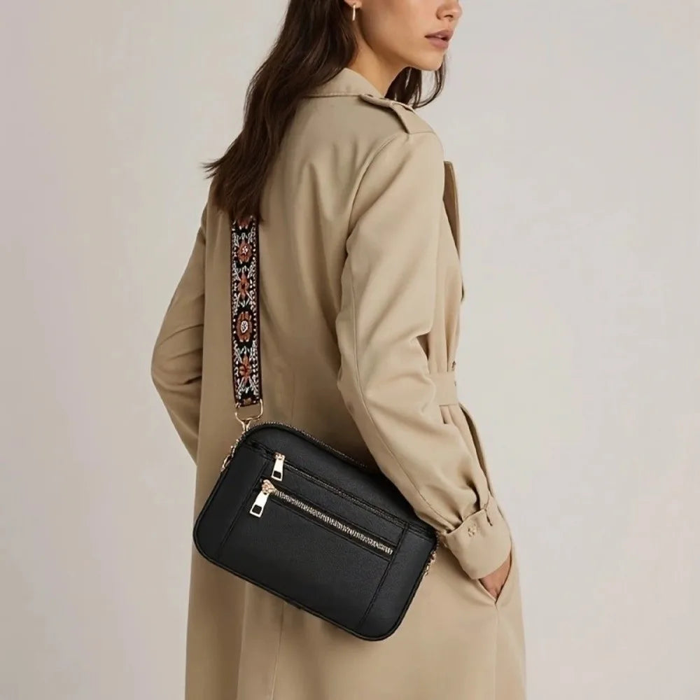 A woman wearing a beige trench coat holds a black retro square crossbody bag with an adjustable strap.