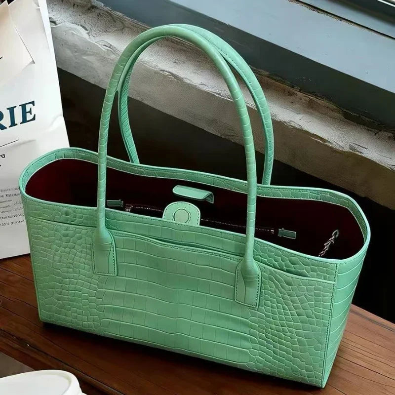 A light green croc embossed leather tote bag with a handle and magnetic closure resting on a wooden surface.