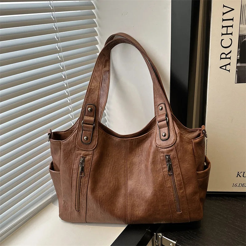 A brown leather everyday large-capacity shoulder tote bag is displayed against a white background next to a window blind.