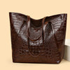A dark brown crocodile print tote bag with leather handles shown in a studio setting.
