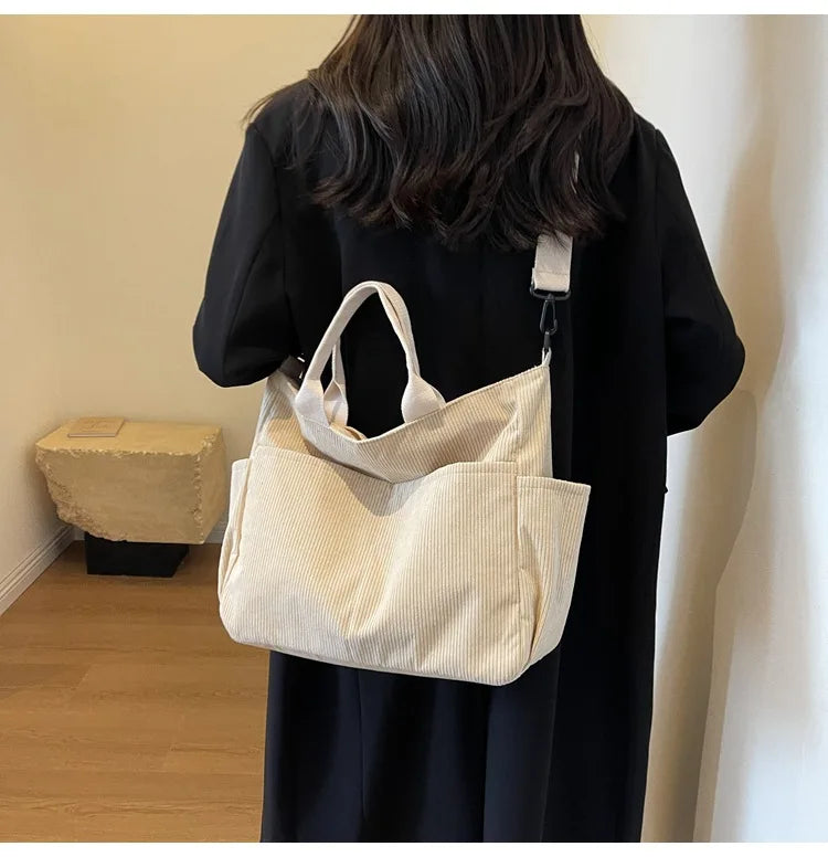 A beige corduroy multi-pocket tote bag is held by a person wearing a black dress.