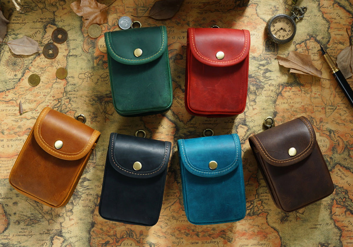 A collection of five leather waist bags in various colors including green, red, brown, black, and blue, arranged on an old ma