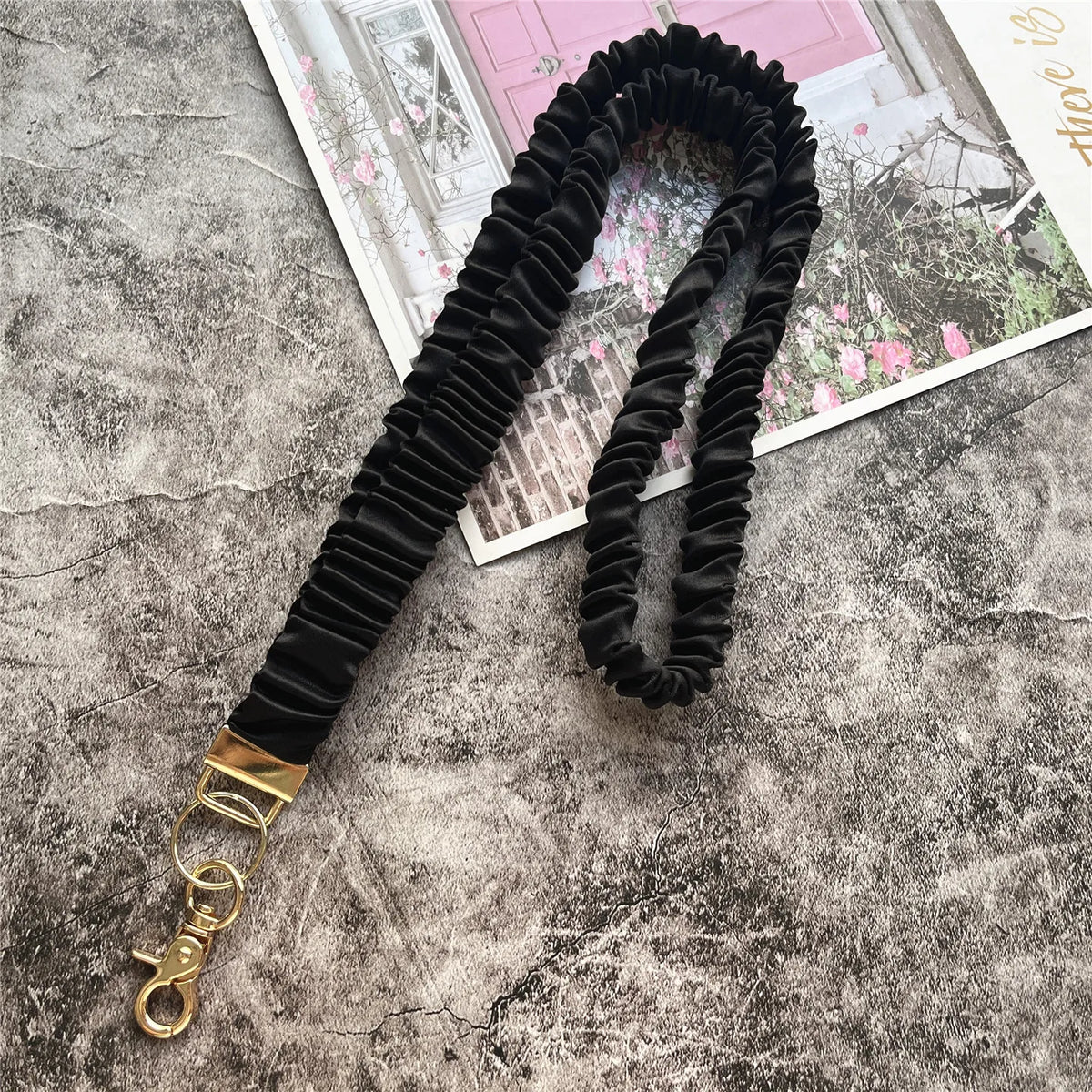 Schwarzes gerafftes Lanyard mit goldenem Schlüsselclip auf einer strukturierten grauen Oberfläche.