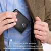 A hand holds a black slim leather wallet against a blue striped shirt and brown jacket.
