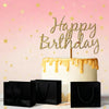 Pastel de cumpleaños blanco con glaseado dorado y una figura que dice Feliz Cumpleaños, junto a bolsas de regalo negras sobre