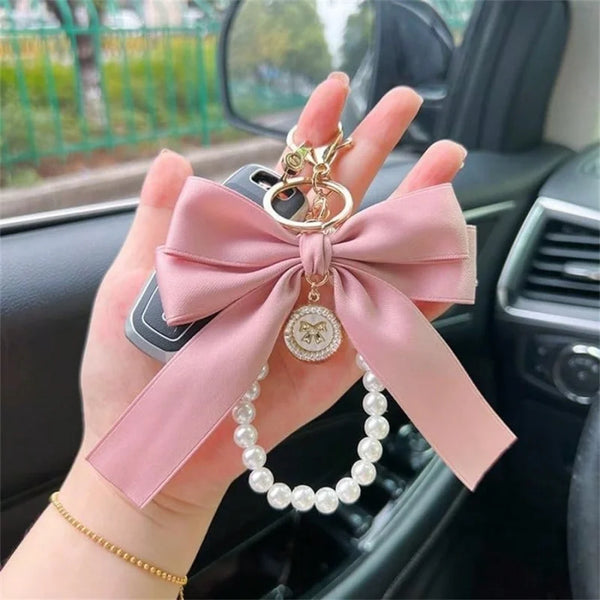 Une main tient un porte-clés nœud papillon rose avec un charm doré et un collier de perles.