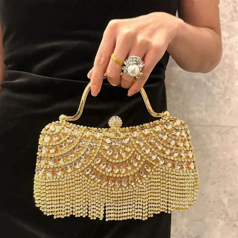 A hand holds an elegant gold and diamond rhinestone evening clutch bag with a golden tassel against a black dress.