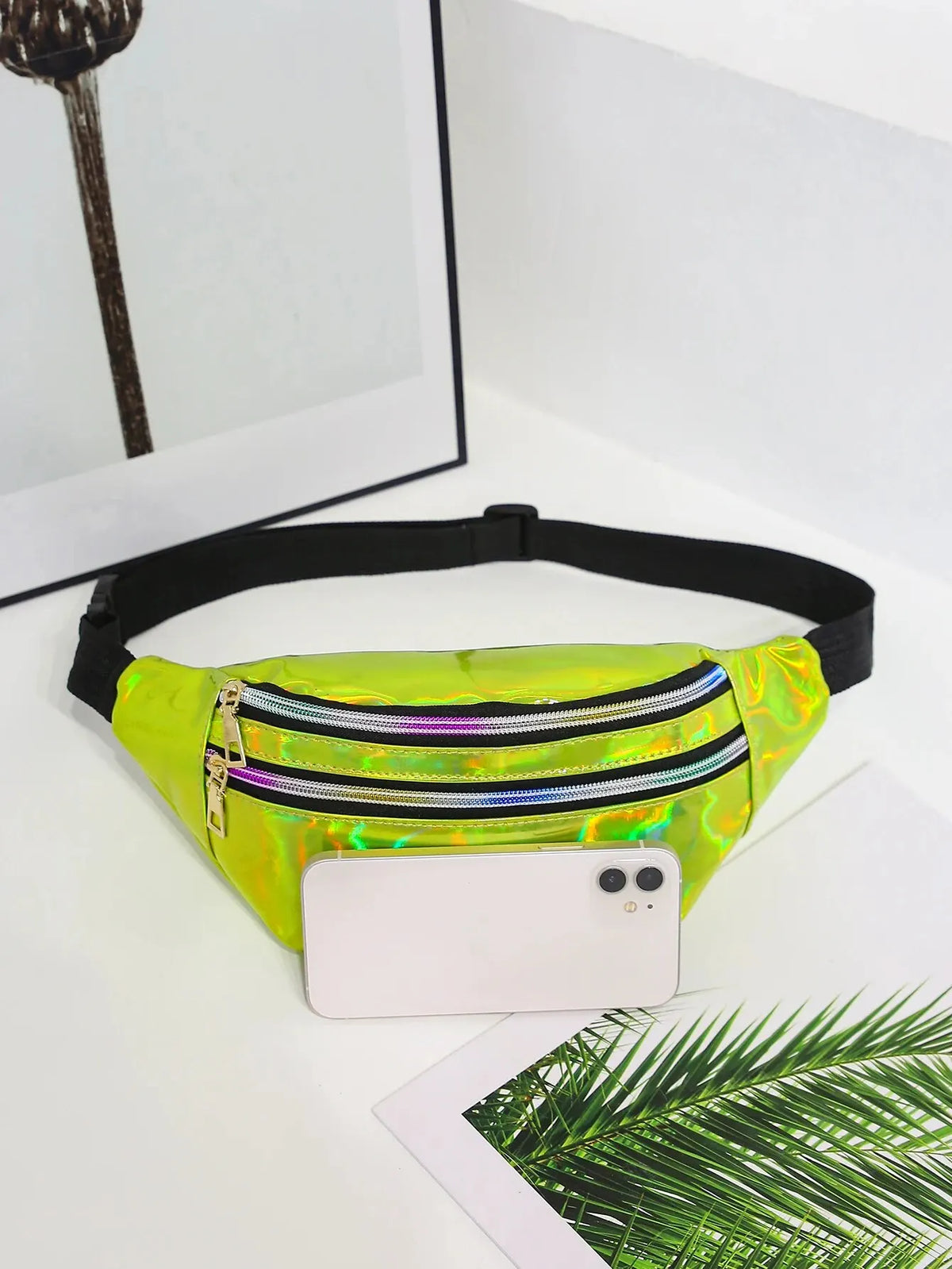 A neon green holographic sequin waist bag with a black adjustable strap resting on a white surface next to a smartphone and p