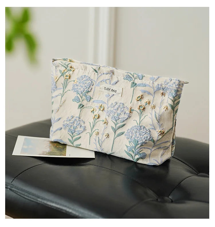 A light blue floral canvas makeup bag with gold embroidery resting on a black leather surface.