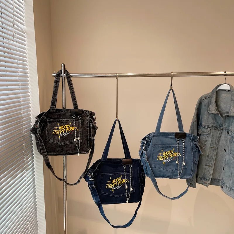 A denim crossbody bag with gold hardware is displayed hanging on a metal rack next to a denim shirt.