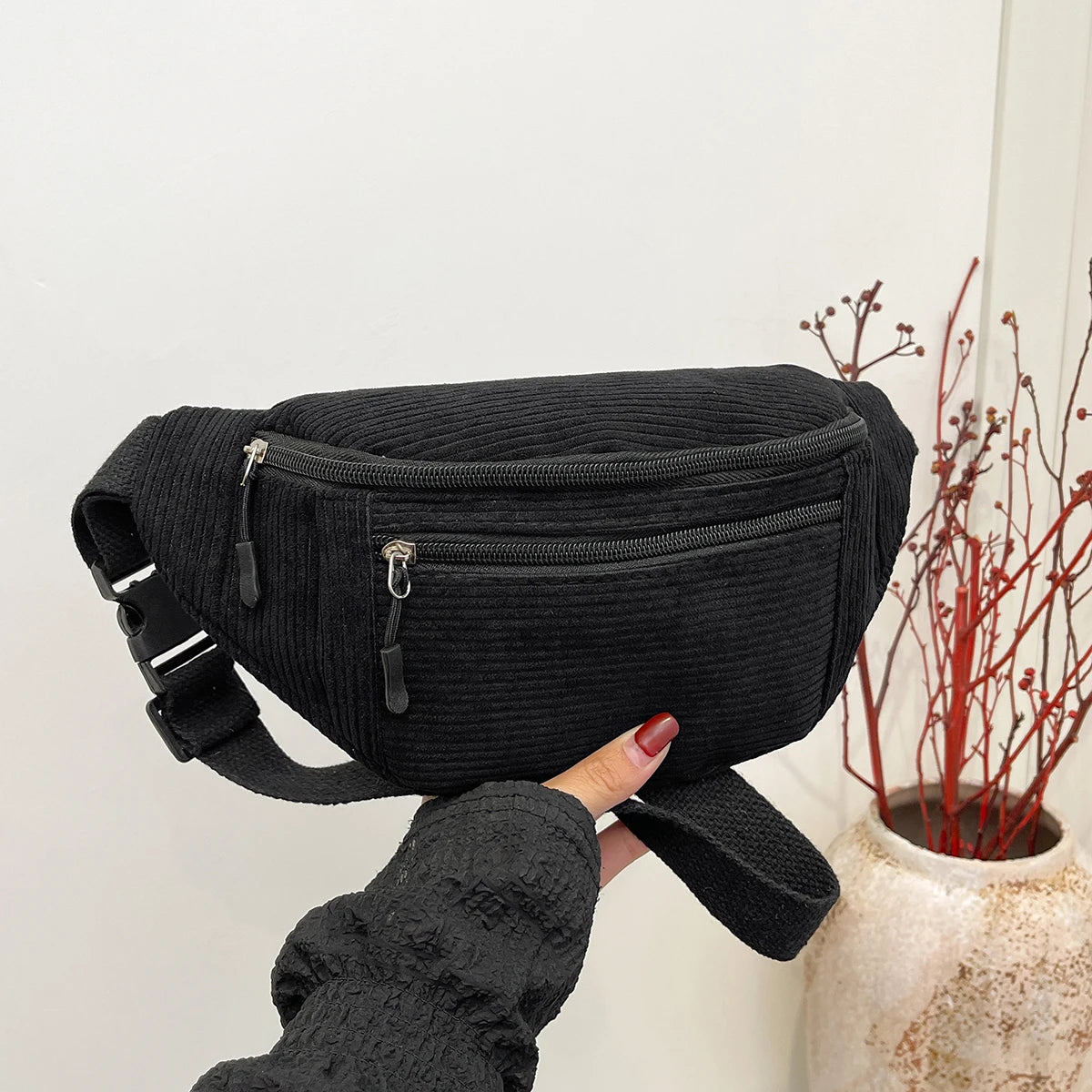 A hand holds a black corduroy crossbody waist bag with an adjustable strap against a white background.
