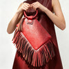 A woman holds a red leather fringed clutch bag with tassels against a light background.