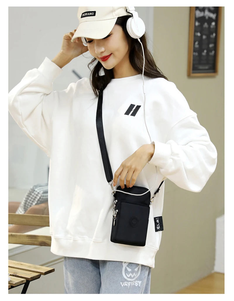 A woman wearing a white sweatshirt and light wash jeans holds a black nylon crossbody phone bag.