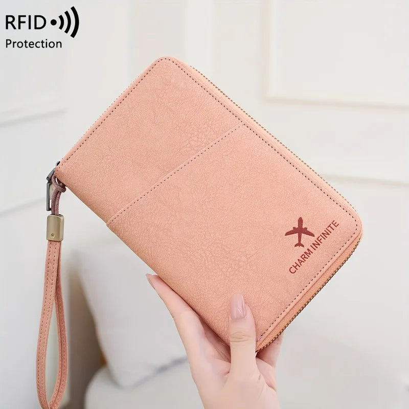 A hand holds a pink leather passport wallet with a wrist strap, featuring an embroidered charm.
