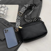A black vegan PU crossbody bag with a patterned strap and a smartphone resting on a gray fabric background.