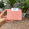 A pink genuine leather zipper card holder and coin purse is held in a hand, set against a backdrop of green plants.