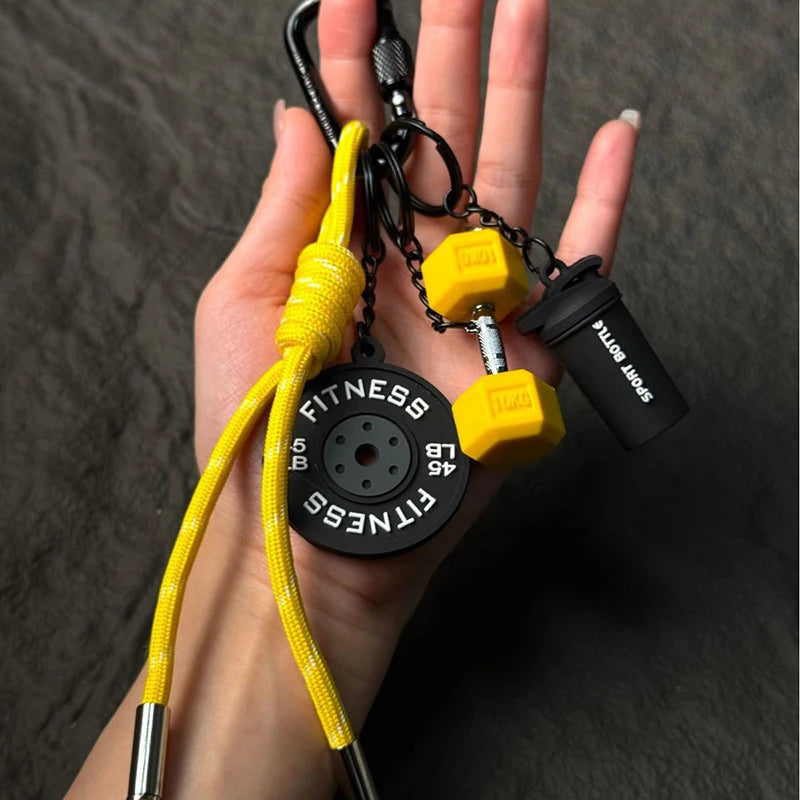 A hand holds a yellow fitness keychain with a black D-shape nylon strap and various gym accessories.