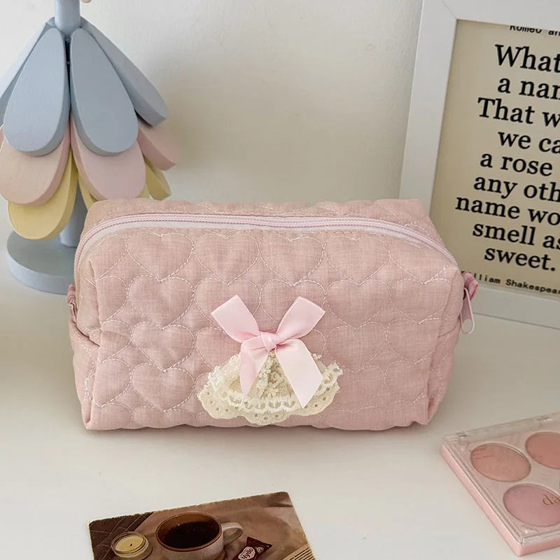 A pink quilted cosmetic storage pouch with a white lace bow resting on a white surface.